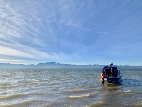 A boat with three people in life jackets bobs on rough water with blue sky and hills in the background.