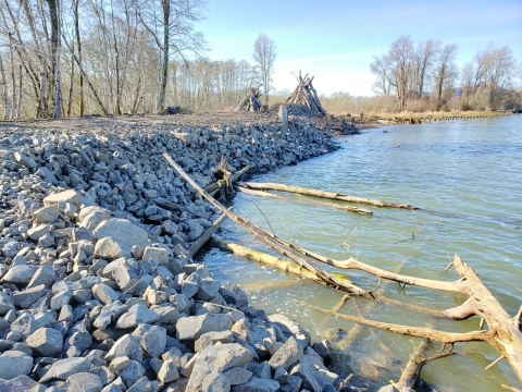 A large wall of rocks next to a river lined with bare trees. A hut built out of sticks is also visible.