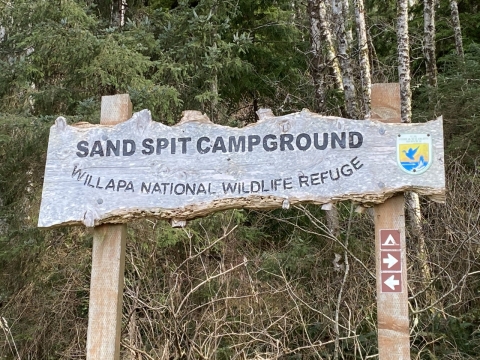 A weathered wooden sign saying "Sand Spit Campground- Willapa National Wildlife Refuge" with an official emblem sticks out of the ground against a background of trees.