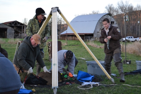 Four people stand in a field around a tripod scale with a hooded and tagged deer.
