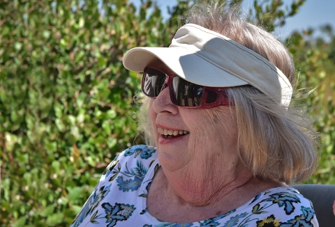 Smiling woman in sunglasses and sun visor sits in chair outside