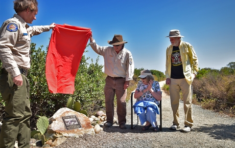 Two men raise red flag in air to reveal a plaque embedded on a rock to woman sitting in chair
