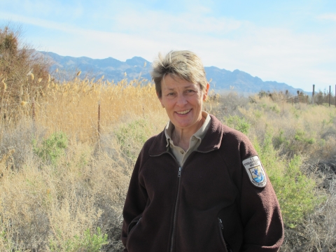 a woman wearing a service jacket stands in front of a desert wetland with mountains in the background