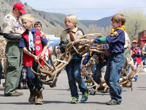 Three young boys who look to be about 10 or 11 years old carry a bunch of elk antlers along a trail with mountains in the backgrounf