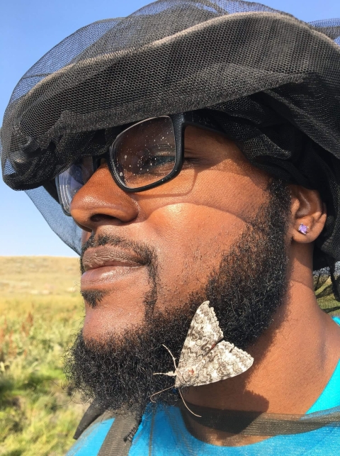 A man in a hat and glasses lets a moth rest on his beard.