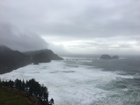 Dense fog and gray clouds hang over the rolling headlands of the pacific coastline, with islands in the distance