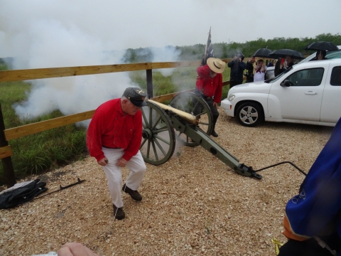 Two men in red shirts turn their backs as an antique cannon fires behind them.