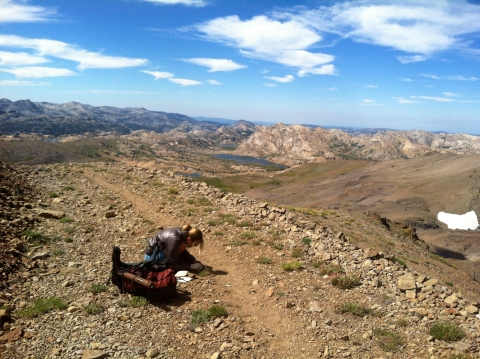 a woman kneels at the edge of a path winding along the ridge of high mountains that meet the blue sky
