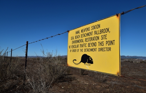 A yellow sign with black lettering hangs on a barbwire fence.