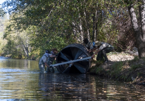 Four people lower a large, round piece of equipment into a stream