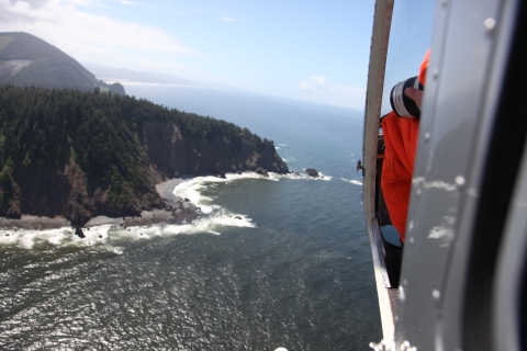 A person in an orange flight suit, holding a camera, sits inside a helicopter far above the Pacific Ocean, with a rolling forested headland in the background