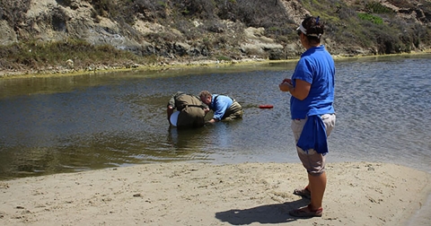 A woman on water's edge watches two men in the creek dip plastic buckets into the water.