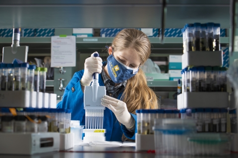 a woman with blonde hair works in a lab wearing a lab coat. She is using a pipette to squeeze samples into test tubes for analysis