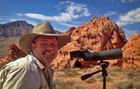 A man with a mustache and goatee wears a khaki shirt and large-brimmed khaki hat out in the desert. He looks through a hunting scope looking for sheep. 