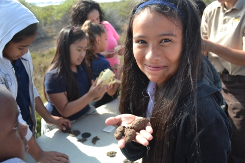 A girl with a funny grin holds fake animal scat at San Diego Bay National Wildlife Refuge. Other students nearby examine other samples.