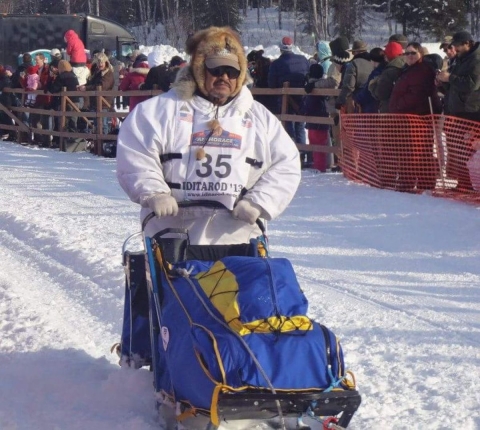 A man in a white puffy jacket, fur-lined hood and sun visor rides a dog sled in the snow past spectators. On his jacket is an Iditarod sign with the number 35.