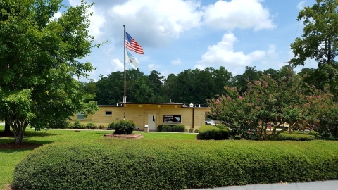 A tan brick building sitting in a grassy courtyard surrounded by trees with a flag pole displaying the American flag and Department of Interior flag.