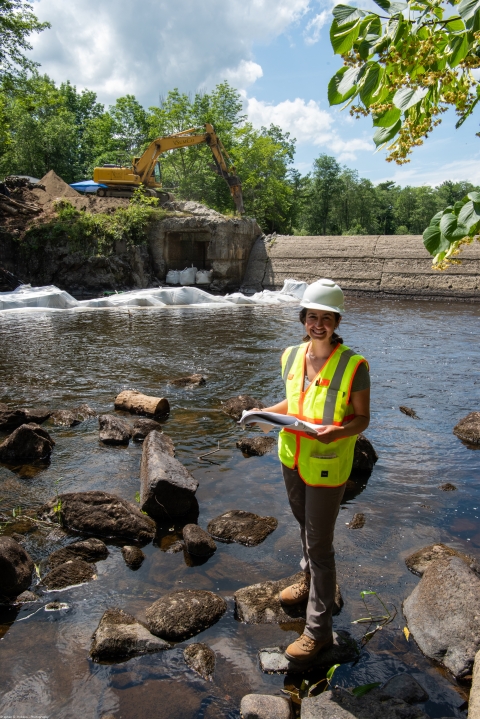 A person in a reflective vest and hard hat stands in a river. 