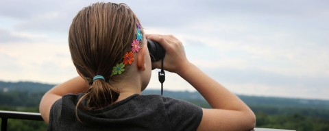 A young woman at a railing looking through binoculars from an overlook