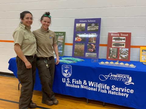 Two biologists (Claudia and Katie) standing in front of a display table at a local school. The table has a blue table cloth that reads" Conserving America's Fisheries, US Fish and Wildlife Service, Nashua National Fish Hatchery". There are several posters of various colors depicting images of American shad, Atlantic salmon, and Brook trout. On the table are many brochures, as well as preserved samples of each of the salmon's life stages.