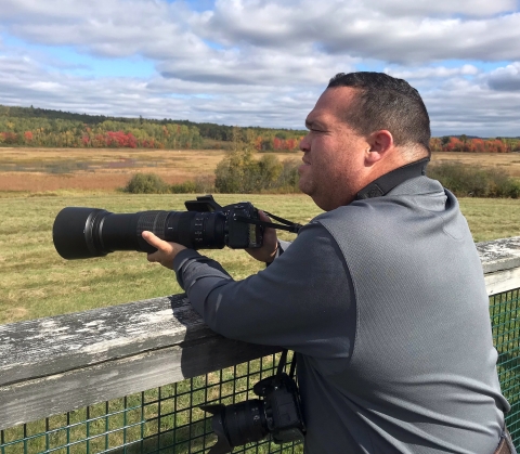 A man holding his camera and leaning against a railing overlooking a meadow and forest
