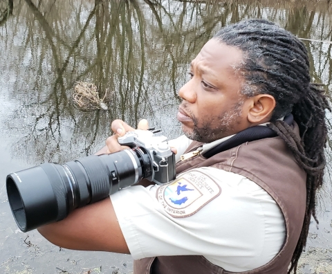 A man with dreadlocks and a beard in a U.S. Fish and Wildlife Service uniform holding a camera near a marsh