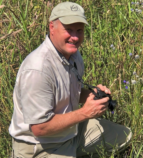 A man wearing a brimmed ballcap crouching down in vegetation and holding his camera