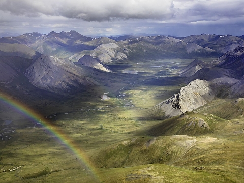 A rainbow over a greening valley in the foreground with snowy mountains in the background