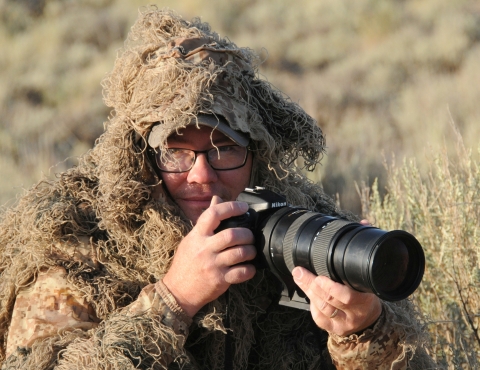 A man with glasses in a camouflage hunting suit holding a camera with a zoom lends in a field from brown grass