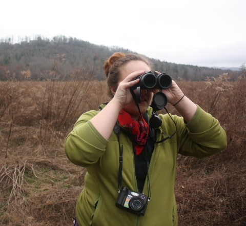 A woman in an olive green top with a camera strung around her neck looks through binoculars at birds.