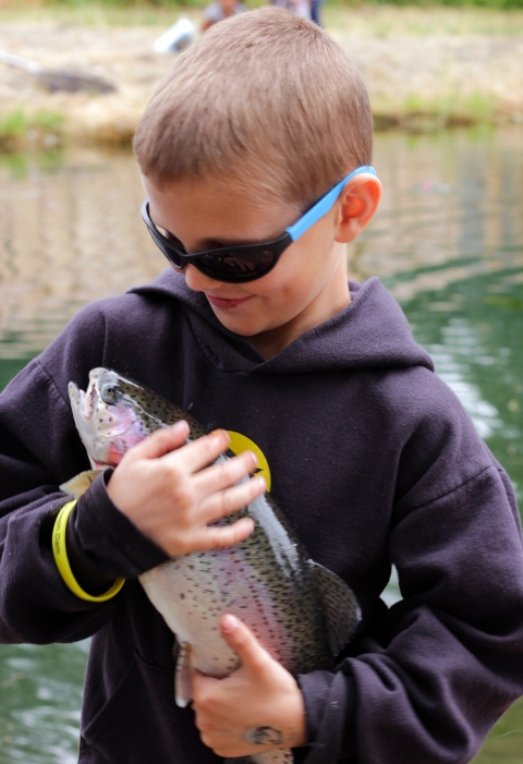 Boy in sweatshirt and sunglasses is pressing a rainbow trout to his chest that is the length of his torso and looking down at it while smiling.