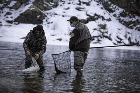 Two men, one in Service uniform, net a fish in a river and prepare to put it into a tube-shaped transport container. Snow is in background, and both men wear coats.