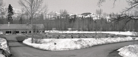 black and white photo of fish ponds in front of the low hatchery building, with snow piled up on either side of the gravel road and on the hills in the background.