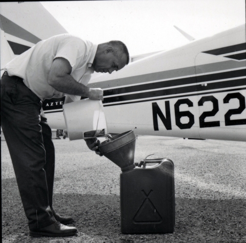 A man in short-sleeved white shirt and dark trousers carefully pours a bucket of fish into a funnel and into a rectangular water container while standing in front of a small airplane.