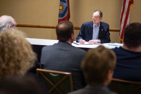 Man seated at a table, signing a document, while a seated crowd looks on