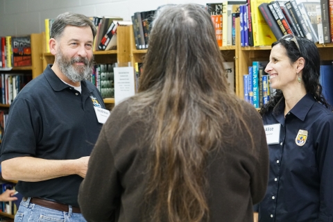 Thee people standing in a circle conversing, with shelves of books in the background