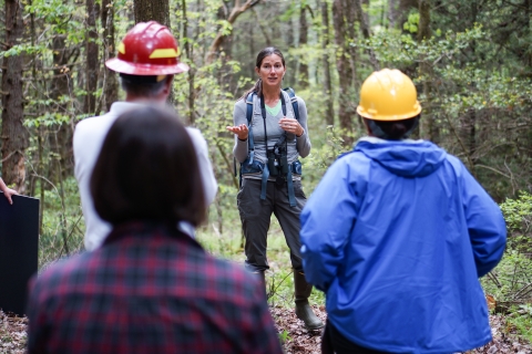 A group of people stands in a forest with one person speaking to the group