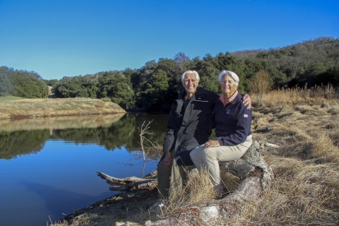 Two elderly people sitting on a log next to a body of water pose for a picture