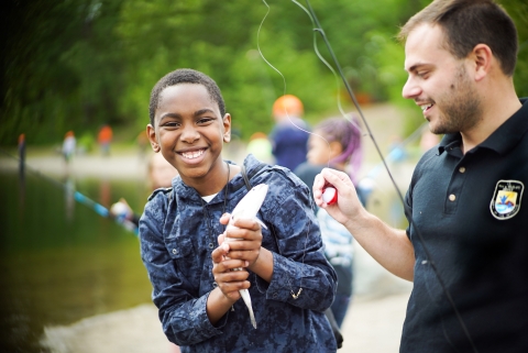 A boy holds a fish in his hands and smiles at the camera. A person next to him holds a fishing rod an bobber. 