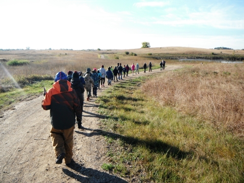 A long single file line of students hiking on the Wetland Way Trail in fall