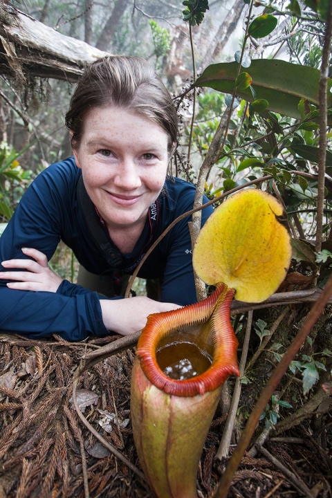 A woman looks smiling into the camera. In front of her is a yellow and red plant that looks like a Venus flytrap.