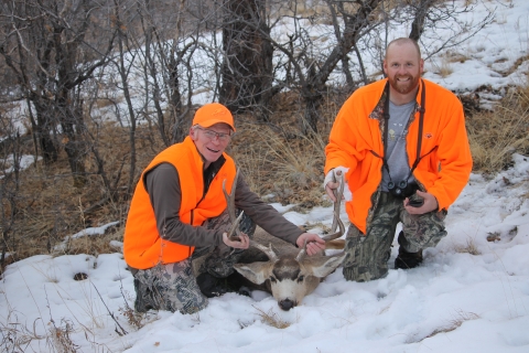 Two men wearing orange crouching next to a harvested mule deer buck in the snow