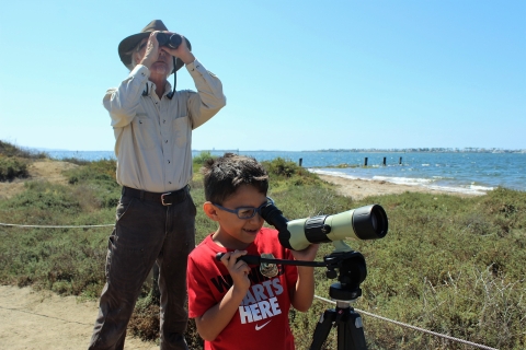 Park ranger using binoculars stands behind a child looking into a scope.