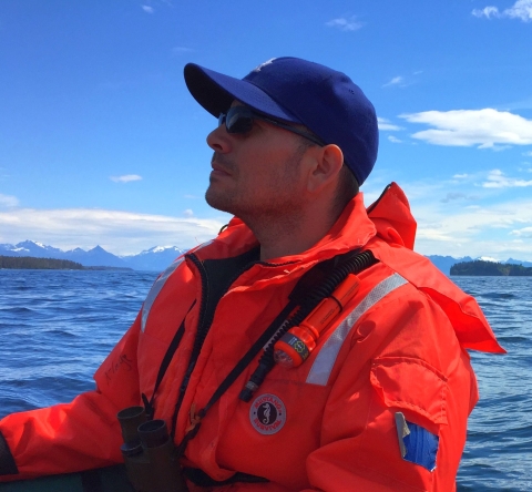 Side view of a man in a blue baseball cap, sunglasses and orange jacket in a boat on water with mountains in the background