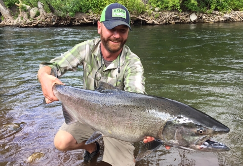 A smiling man in shorts and ballcap kneels at the edge of a river with a huge summer chinook salmon held out to the camera.