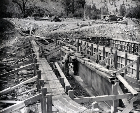 Black and white photo of worker in 1941 removes wooden molds from newly poured concrete, long, oval fish ponds, old cars parked in background in gravel area.