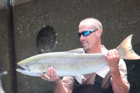 A man in waders and sunglasses holds up a large salmon horizontally in both hands.