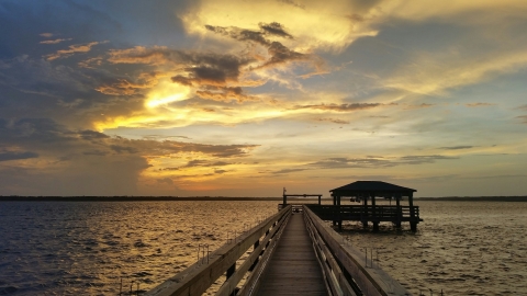 the sun shinning through the clouds as it sets over the water with the dock to the boat lift in the foreground