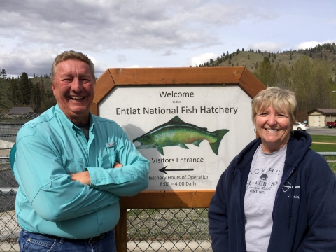 A smiling man and woman flank a small sign reading "Welcome to Entiat National Fish Hatchery."