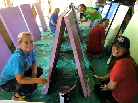 Teenage girls in t-shirts paint folding wooden signboards on a tarp while smiling at the camera.